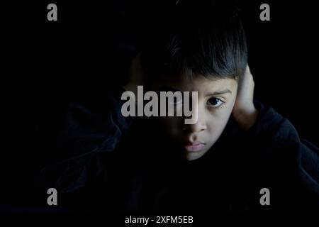 latino boy in the dark staring at the camera angrily covering his ears because he is being bullied Stock Photo