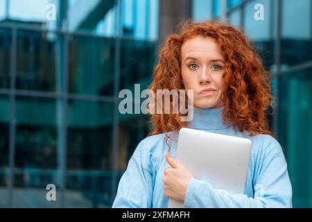 worried young woman graduate with curly red hair biting her lip, holding laptop and excited  about news regarding her job application Stock Photo