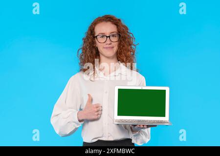 happy redhead woman with glasses  in white shirt showing empty laptop screen, giving thumb up, with positive expression,    on blue background. Mobile Stock Photo