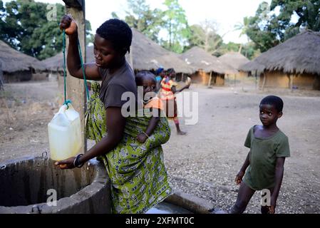 Mother with baby on back collecting water from well, boy observing them, Formosa Island, Bijagos UNESCO Biosphere Reserve, Guinea Bissau, February 2015. Stock Photo