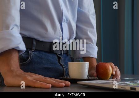 Man gets up to take a break during his workday and has a cup of coffee and a piece of fruit in front of his laptop dressed casually Stock Photo