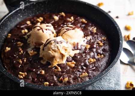 Homemade brownie cooked in a skillet, not baked, with melting vanilla ice cream scoops, nuts, and chocolate chips on a white table Stock Photo