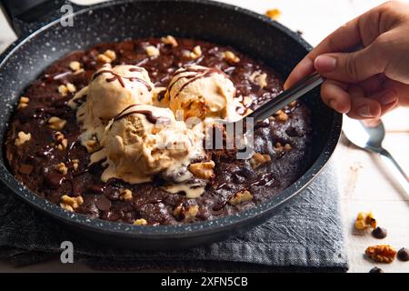 Homemade brownie cooked in a skillet, not baked, with melting vanilla ice cream scoops, nuts, and chocolate chips and a womans hand taking a piece wit Stock Photo