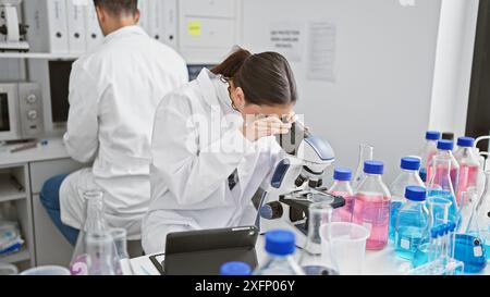 A woman scientist using a microscope in a laboratory with a male coworker and various chemical bottles in the background. Stock Photo