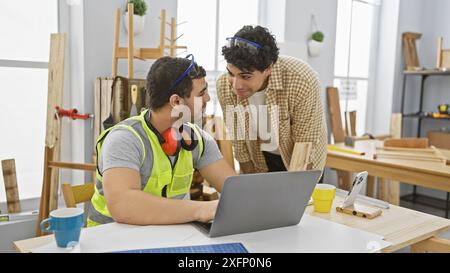 Two men collaborating in a bright carpentry workshop with one using a laptop and the other observing Stock Photo