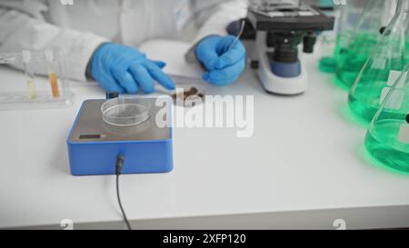 A scientist wearing gloves handling samples in a laboratory equipped with a microscope and beakers of green liquid. Stock Photo