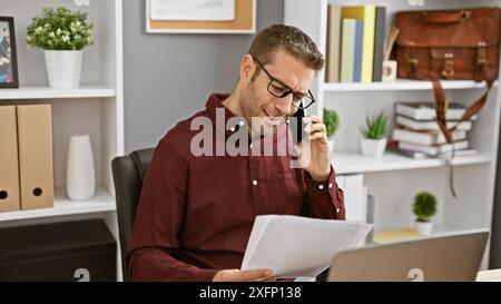 Smiling hispanic man in an office engaging in business over the phone while reviewing documents. Stock Photo