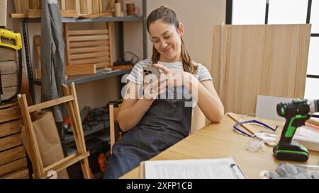 A smiling young woman in a denim apron uses a smartphone while taking a break in a bright carpentry workshop. Stock Photo
