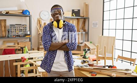Smiling, confident black man with crossed arms wearing safety glasses and headphones in a well-equipped carpentry workshop. Stock Photo