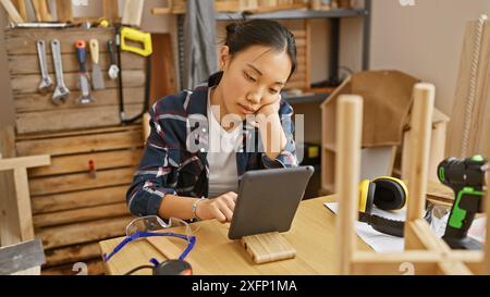 A focused asian woman using a tablet in a cluttered carpentry workshop surrounded by tools and wood. Stock Photo