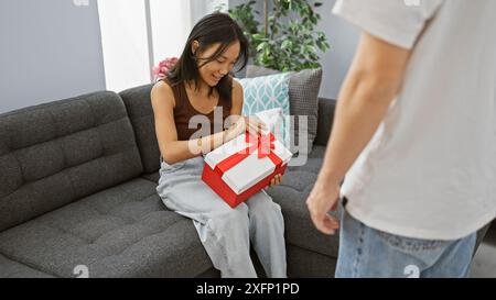 A woman seated on a gray couch receives a red-gift from a man in a cozy living room setting, expressing happiness and surprise. Stock Photo