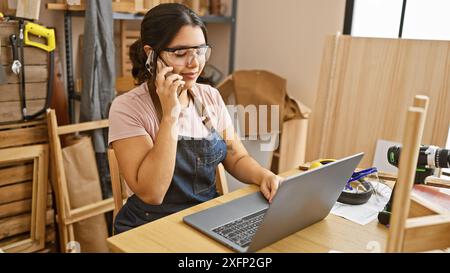 Hispanic woman in a carpentry workshop communicating on phone while using laptop. Stock Photo