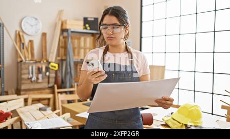 A young hispanic woman examines plans while multitasking with a phone in a well-equipped carpentry workshop. Stock Photo