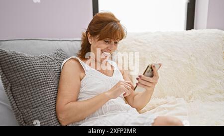 A smiling mature woman enjoys browsing her smartphone while relaxing on a cozy sofa in a bright home interior. Stock Photo