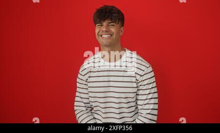 A handsome young hispanic man in a striped shirt smiles against an isolated red background wall, showcasing his attractive features in this vibrant po Stock Photo
