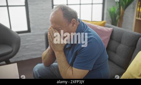 A distressed senior man sits indoors at home with hands covering face, exhibiting stress or sadness. Stock Photo