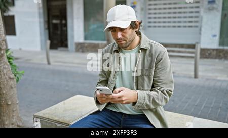 A young man with a beard wearing a cap and jacket using a smartphone while sitting on a bench on an urban city street. Stock Photo