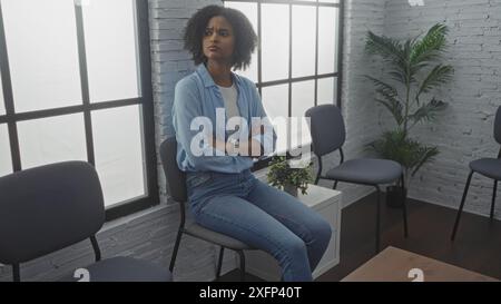 A young african american woman with curly hair and her arms crossed sits in an indoor waiting room with chairs and plants, wearing a blue shirt and je Stock Photo