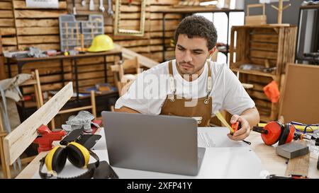 A focused young man with a beard in a carpentry workshop using a laptop and measuring tape. Stock Photo