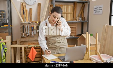 Hispanic woman multitasking in a carpentry workshop, conversing on the phone and writing notes next to a laptop. Stock Photo