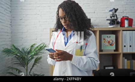 A young african american female doctor with curly hair stands in a hospital room, looking at her phone, wearing a white lab coat and stethoscope. Stock Photo