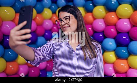 Young beautiful hispanic woman visiting colorful futuristic exhibition space taking selfie Stock Photo