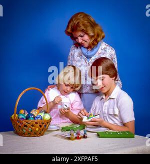 Mother and two children decorating colorful Easter eggs at table with basket, paint and tools, smiling family moment against bright blue studio backgr Stock Photo