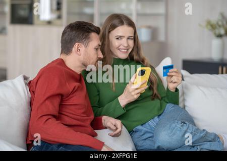Happy married couple, spouses on couch with phone, bank credit card shopping together online. Stock Photo