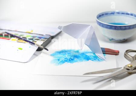 Folded origami dolphin on paper with painted water, crafting a homemade greeting card with scissors, brush and watercolor box, copy space, selected fo Stock Photo