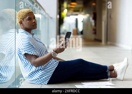 Sitting on floor, woman holding smartphone and looking thoughtful in office hallway Stock Photo