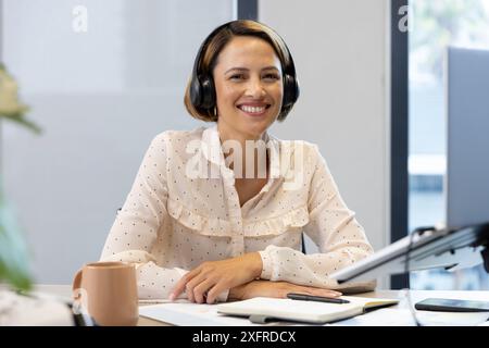 Smiling woman wearing headphones, working at desk with laptop and coffee mug Stock Photo