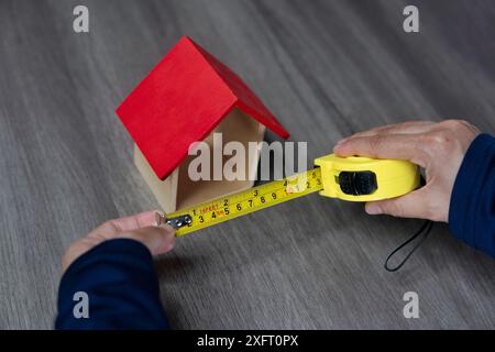 A close-up image of a person holding a yellow tape measure against a miniature wooden house. Home improvement and renovation concept. Stock Photo
