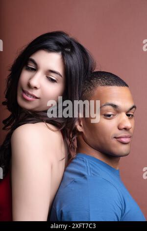 Young couple is posing back to back, with the woman slightly leaning on the man's back, both showing different expressions Stock Photo