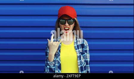 Stylish crazy young woman in glasses, cool modern teenage girl in red baseball cap posing on blue wall background Stock Photo