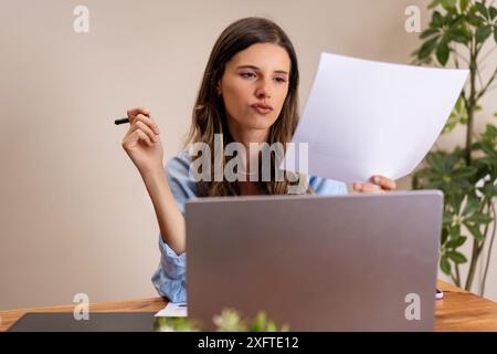 A focused woman reviewing documents at her desk in a home office. Professional and concentrated - remote work, document analysis, modern home office, Stock Photo