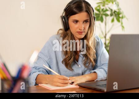 A focused woman wearing headphones and taking notes while working on a laptop. Home office environment - professional remote work, online study sessio Stock Photo