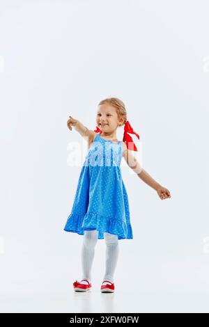 Little beautiful girl, child in cute blue dress pointing somewhere with cheerful smile isolated on white background. Curiosity in childhood Stock Photo