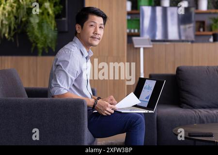 Working on laptop, man analyzing financial charts and holding documents in office Stock Photo