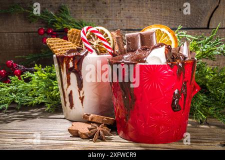 Christmas styled overloaded hot chocolate mugs with various toppings – chocolate slices, spices, sweets, cookies, candy and gingerbreads, wooden cozy Stock Photo