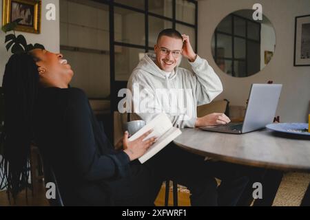 Happy freelancer sitting with girlfriend laughing at home office Stock Photo