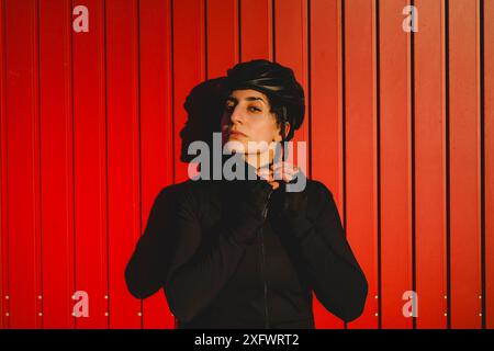 Confident female cyclist putting on helmet against red striped wall Stock Photo