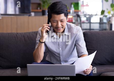 Talking on phone, man holding documents and using laptop while working from home Stock Photo