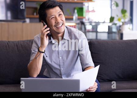 Smiling man holding paper and talking on smartphone, working on laptop Stock Photo