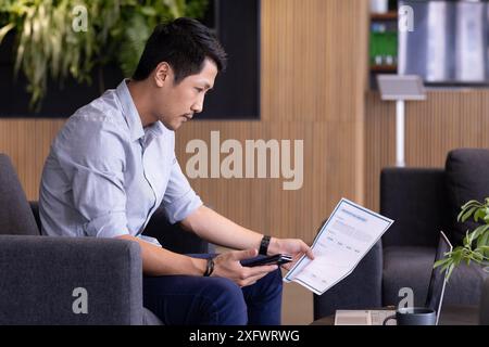 Reviewing financial documents, man holding smartphone and sitting in modern office Stock Photo
