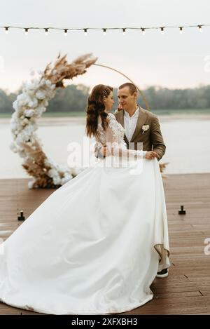 the first wedding dance of the bride and groom on the pier near the river, against the backdrop of the wedding arch Stock Photo
