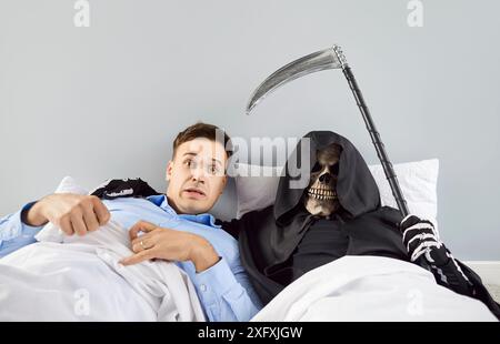 Shocked scared man lying on bed with skeleton in a grim reaper death costume holding scythe Stock Photo
