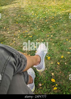 Women's legs in casual wear and white sneakers on lawn with fall leaves Stock Photo