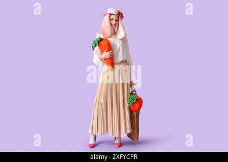 Stylish woman with bunny ears holding carrot toys on lilac background. Easter celebration Stock Photo