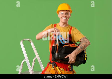 Young male electrician on stepladder with tool bag against green background Stock Photo