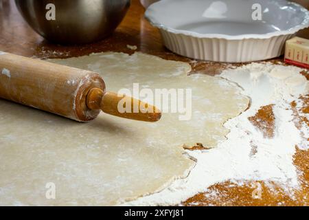Making a pie.   Dough on a floured wooden surface.  Side view includes rolling pin, prepared sliced apples, white pie plate, butter and a wooden rolli Stock Photo